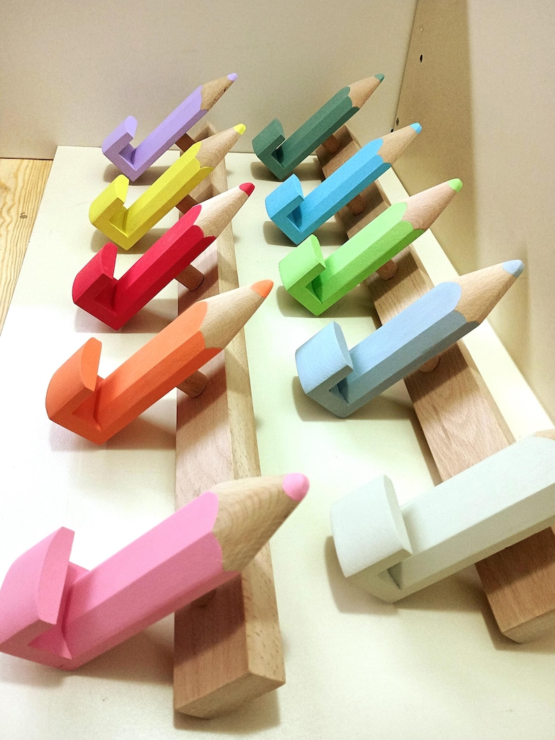 May include: A set of colorful pencil-shaped hooks mounted on a wooden base. The hooks are painted in various colors, including purple, yellow, red, orange, green, blue, light blue, pink, and white. The hooks are designed to resemble pencils with a wooden body and a colored tip.