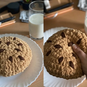 Könnte beinhalten: Ein gehäkeltes Plüschtier in Form eines Schokoladen-Chip-Kekses mit braunen Schokoladenstückchen. Der Keks liegt auf einem weißen Teller.