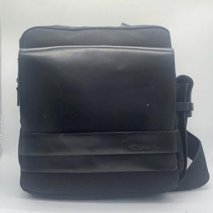 May include: A black messenger bag featuring a smooth, rectangular front flap made of a matte material. A horizontal strap crosses the lower section of the flap, embossed with the Calvin Klein logo. The bag includes a wide, adjustable shoulder strap.