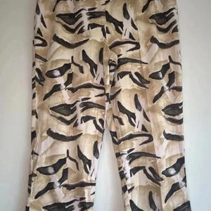 Puede incluir: Pantalones de talle alto con fondo beige y crema y un patrón abstracto de rayas de tigre en marrón oscuro. Los pantalones incluyen una cinturilla con trabillas, cierre de botón frontal y pequeñas aberturas en el bajo de cada pernera.