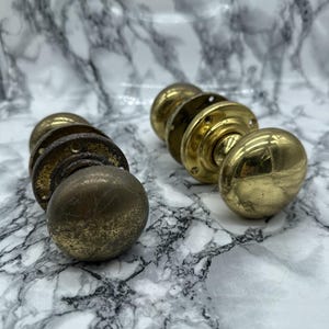 May include: Two round brass door knobs with circular mounting plates. One knob has a polished, reflective gold-tone finish, while the other knob features a weathered, tarnished, and darkened patina with visible surface texture and oxidation.
