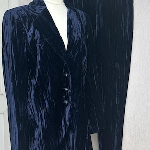 May include: A dark navy blue two-piece set consisting of a long-sleeved blazer and matching trousers. The blazer features a notched lapel, three front buttons, and a crushed velvet texture. The trousers are straight-legged with a matching finish.