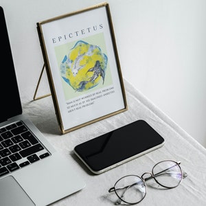 May include: A gold-framed print with a quote by Epictetus, "Man is not worried by real problems so much as by his imagined anxieties about real problems." The print features a yellow background with three black and white fish swimming in a circle. The print is sitting on a white surface with a laptop, a black smartphone, and a pair of round glasses.
