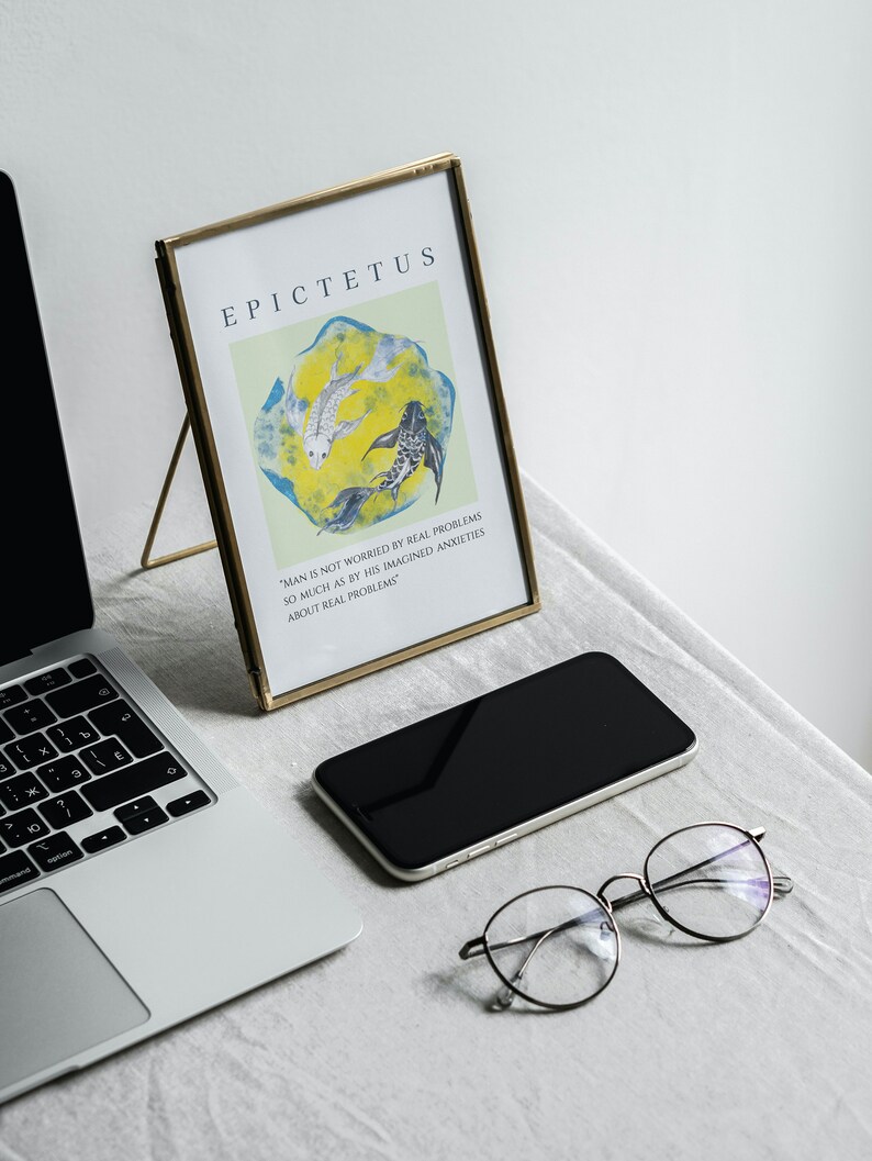 May include: A gold-framed print with a quote by Epictetus, "Man is not worried by real problems so much as by his imagined anxieties about real problems." The print features a yellow background with three black and white fish swimming in a circle. The print is sitting on a white surface with a laptop, a black smartphone, and a pair of round glasses.