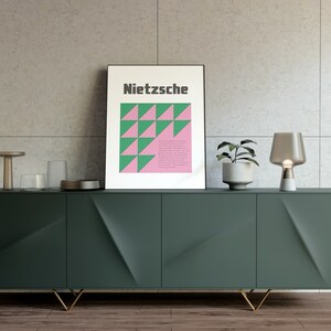 May include: A dark green cabinet with gold legs and three doors. The cabinet is decorated with a geometric pattern. A framed poster with the word "Nietzsche" and a geometric pattern in pink and green is on top of the cabinet. A small table lamp with a clear glass shade and a white base is on the right side of the cabinet. A small potted plant is on the left side of the cabinet.