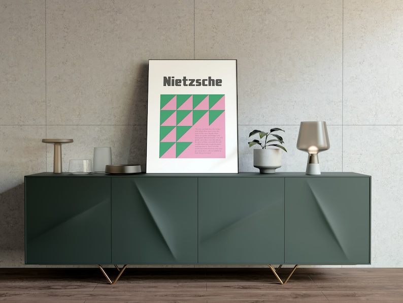 May include: A dark green cabinet with gold legs and three doors. The cabinet is decorated with a geometric pattern. A framed poster with the word "Nietzsche" and a geometric pattern in pink and green is on top of the cabinet. A small table lamp with a clear glass shade and a white base is on the right side of the cabinet. A small potted plant is on the left side of the cabinet.
