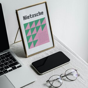May include: A gold-framed picture with a pink and green geometric design and the word "Nietzsche" in black text. A laptop, a smartphone, and a pair of glasses are also on the table.