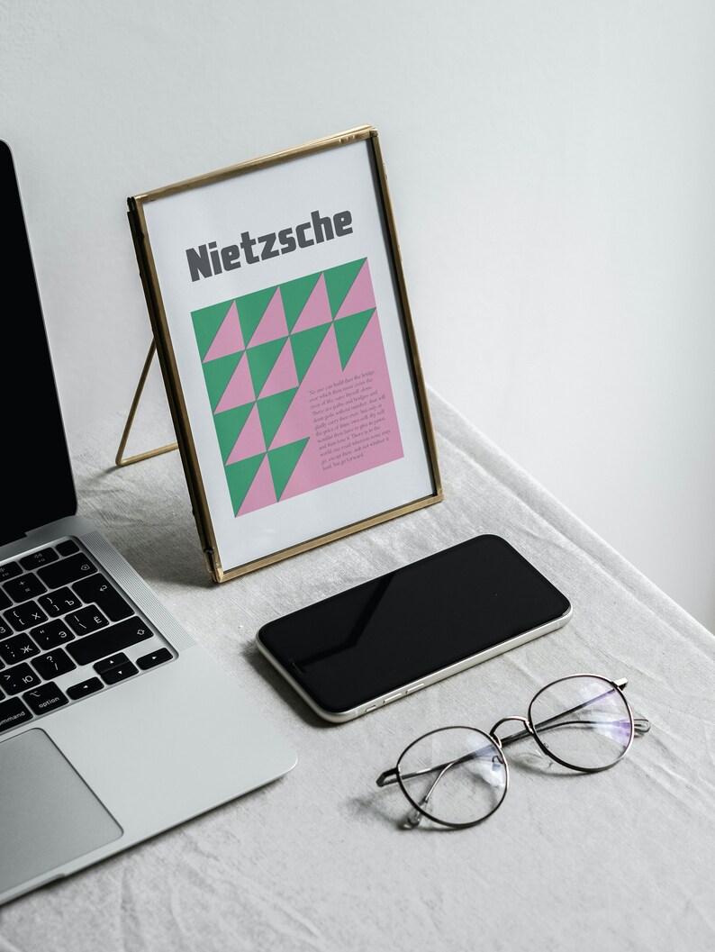 May include: A gold-framed picture with a pink and green geometric design and the word "Nietzsche" in black text. A laptop, a smartphone, and a pair of glasses are also on the table.