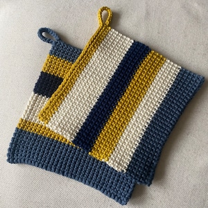 Puede incluir: Dos agarraderas de olla de ganchillo con un patrón a rayas en blanco, azul marino y amarillo mostaza. Las agarraderas están hechas con un punto texturizado.