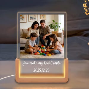 以下が含まれることがあります： 家族写真が入った長方形の照明付きフォトフレーム。フレームは温かみのあるピーチ色のベースと光るボーダーが特徴です。フレームには「You make my heart smile」と「2025.12.26」の文字が書かれています。写真は4人家族です。