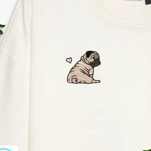 Könnte beinhalten: Cremefarbenes Sweatshirt mit einem gestickten Mops und einem rosa Herz. Der Mops ist in sitzender Position dargestellt, mit detaillierten Schattierungen und einem konzentrierten Ausdruck. Das Design befindet sich auf der linken Brustseite des Kleidungsstücks.