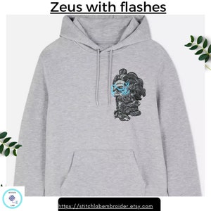 Puede incluir: Una sudadera con capucha gris claro con un bolsillo delantero y una capucha con cordón. La sudadera presenta un diseño bordado de una figura con una calavera y rayos azules. El texto "Zeus with flashes" está encima del diseño.