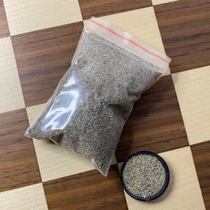 Peut inclure: Un sac en plastique transparent rempli d'une substance granulaire brune et grise. Un petit récipient noir contenant la même substance se trouve sur une surface à carreaux brun et blanc.
