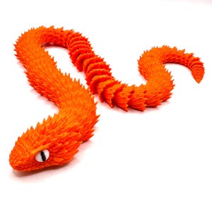 Op de afbeelding: Een oranje, gelede slangenknuffel met een textuur, schubachtig ontwerp. De slang heeft een gedetailleerde kop met een enkel oog en een gesegmenteerd lichaam dat flexibele beweging mogelijk maakt. Het speelgoed staat tegen een witte achtergrond.