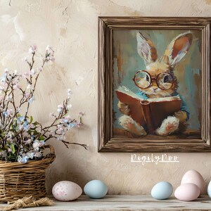 Puede incluir: Una pintura enmarcada de un conejo marr&oacute;n con gafas leyendo un libro. El conejo est&aacute; sentado en una manta azul y blanca. La pintura est&aacute; en un marco de madera marr&oacute;n con un paspart&uacute; blanco. La pintura est&aacute; colgada en una pared beige. Hay cinco huevos de Pascua de color pastel en un estante de madera debajo de la pintura.
