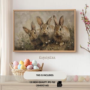 May include: A painting of three brown rabbits with white fur on their chests. The rabbits are sitting in a field of green grass and white flowers. The painting is framed in a light brown wood frame.