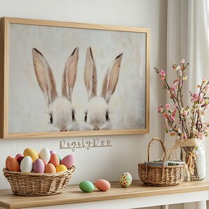 May include: A framed painting of two white bunnies with pink noses peeking out from behind a white background. The painting is hanging on a white wall above a table with a basket of colorful Easter eggs and a vase of pink flowers.