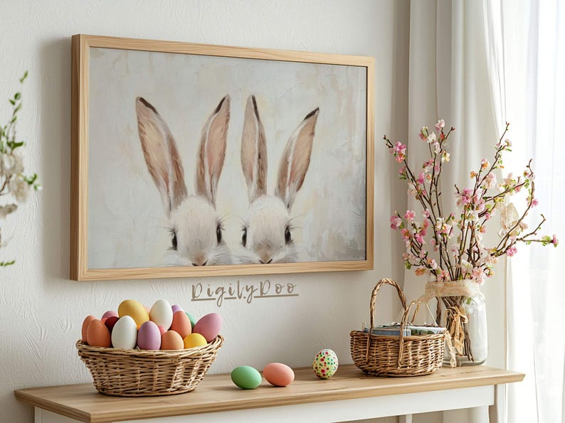 May include: A framed painting of two white bunnies with pink noses peeking out from behind a white background. The painting is hanging on a white wall above a table with a basket of colorful Easter eggs and a vase of pink flowers.