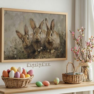 May include: A framed painting of three brown rabbits sitting in a field of green grass and white flowers. The painting is in a light wood frame and is hanging on a white wall.