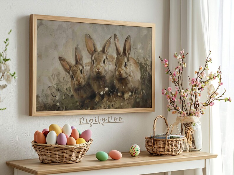 May include: A framed painting of three brown rabbits sitting in a field of green grass and white flowers. The painting is in a light wood frame and is hanging on a white wall.