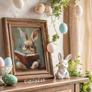 Puede incluir: Una pintura enmarcada de un conejito con gafas leyendo un libro, con huevos de Pascua de colores pastel colgando de una vid en el fondo. Una figurita de conejito de cer&aacute;mica blanca se sienta sobre una mesa de madera.