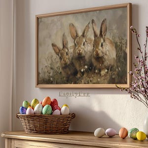 May include: A framed painting of three brown rabbits with long ears, set on a wooden table with a basket of colorful Easter eggs and a vase of pink flowers.