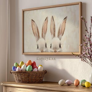 May include: A framed painting of two white bunnies with brown ears and noses. The bunnies are looking at the viewer. The painting is on a white wall with a vase of pink flowers and a basket of colorful Easter eggs on a wooden table.