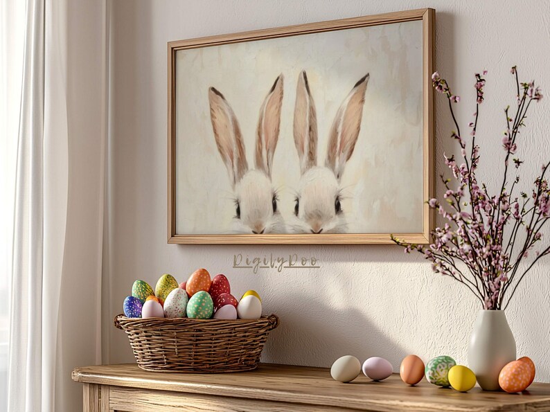 May include: A framed painting of two white bunnies with brown ears and noses. The bunnies are looking at the viewer. The painting is on a white wall with a vase of pink flowers and a basket of colorful Easter eggs on a wooden table.