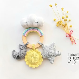 Könnte beinhalten: Ein gehäkeltes Babyrassel mit einem Regenbogen, einer Wolke, einem Stern und einer Sonne. Die Rassel ist aus einem Holzring gefertigt und hat einen pastellfarbenen Regenbogen, der um ihn herum gehäkelt ist. Die Wolke ist weiß, der Stern ist grau und die Sonne ist gelb.