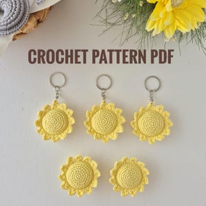 Può includere: Portachiavi e applicazioni a forma di girasole all'uncinetto giallo. I portachiavi hanno un anello e una catena argentati. Le applicazioni e i portachiavi sono realizzati in filato giallo. Il testo "CROCHET PATTERN PDF" è nella parte superiore dell'immagine.