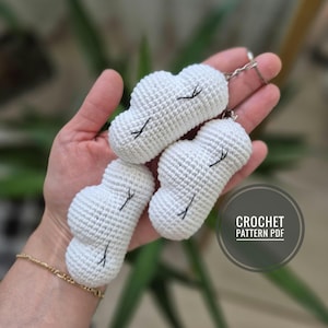Puede incluir: Tres llaveros de nube blancos de ganchillo con pestañas cosidas en negro. Los llaveros se sostienen en una mano. La imagen también incluye un gráfico circular con el texto "CROCHET PATTERN PDF".