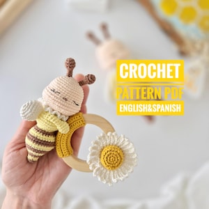 May include: A handmade crochet baby rattle shaped like a bee, featuring a light tan head, yellow and brown striped body, and a wooden ring with a white daisy. The text "CROCHET PATTERN PDF ENGLISH&SPANISH" is visible.