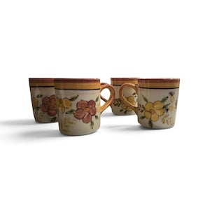 May include: A set of four ceramic mugs featuring a hand-painted floral pattern with yellow and reddish-brown flowers on a cream background. Each mug has a horizontal orange and yellow band near the rim and a curved handle on the side.