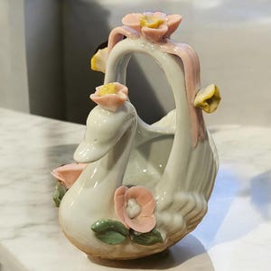 Puede incluir: Una figurita de cisne de porcelana blanca con flores rosas y amarillas. El cisne sostiene una pequeña cesta con un diseño floral.
