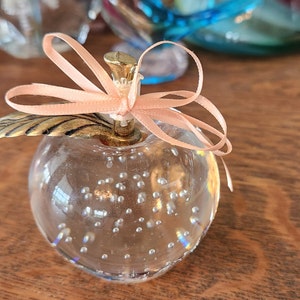 Op de afbeelding: Een heldere glazen appel met een gouden blad en een roze strik. De appel is gevuld met kleine luchtbellen.