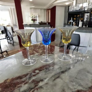 Op de afbeelding: Drie glazen shotglazen met verschillende gekleurde glazen bodems. De glazen zijn helder en hebben een klokvormige kom. De bodems zijn geel, blauw en helder.