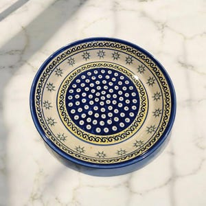 Può includere: Una ciotola rotonda in ceramica con bordo blu e interno color crema. La ciotola presenta un motivo a pois blu e bianchi al centro, circondato da un bordo decorativo giallo e blu. Il bordo esterno ha un motivo a stelle ripetute.