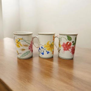 Op de afbeelding: Drie witte keramische mokken met bloemmotieven. Elke mok heeft een ander bloemmotief in geel, roze en rood, met groene accenten. De mokken hebben een zwarte rand en witte handvatten, geplaatst op een houten oppervlak.