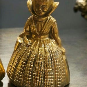 May include: A gold-colored metal bell shaped like a girl wearing a dress and a bonnet. The bell has a detailed design and is a decorative item.