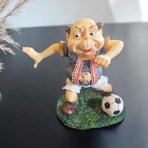 May include: A figurine of a man playing soccer. He is wearing a blue and white jersey with pink trim, white shorts, and brown shoes. He is kicking a black and white soccer ball on a green grass base.
