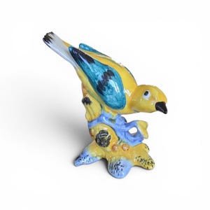 Puede incluir: Figura de pájaro de cerámica posada en una rama. El pájaro tiene plumaje amarillo con detalles azules y negros en las alas y la cola. La rama es amarilla y azul con una base negra y marrón. El pájaro tiene un pico negro.