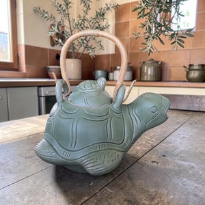 Może przedstawiać: Zielony ceramiczny czajnik w kształcie żółwia, z bambusowym uchwytem. Czajnik ma szczegółowy wzór skorupy żółwia i małą figurkę żółwia na pokrywce. Czajnik stoi na drewnianej powierzchni.