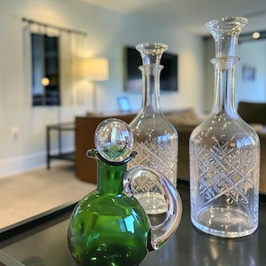 Puede incluir: Una colección de botellas y decantadores de vidrio decorativos. El primer plano presenta una botella de vidrio verde con asa y tapón transparente. Detrás hay dos decantadores de vidrio transparente con diseños grabados. Las botellas están sobre una superficie oscura.