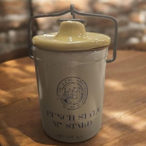 May include: A white ceramic jar with a yellow lid and a metal wire handle. The jar has a blue label that reads "Pinch St. Vt. Stand" and a blue circular logo with the text "Ro. Lingst. J. Zac. Ary. Minnesota. N.E.K."