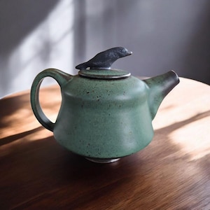 以下が含まれることがあります： 鳥の形をした蓋が付いた緑色の陶器製のティーポット。ティーポットは斑点のある釉薬と丸みを帯びたボディをしています。
