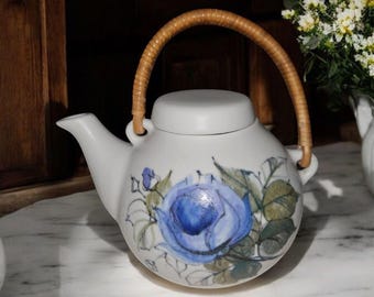 May include: White ceramic teapot with a blue rose design and a brown wicker handle.