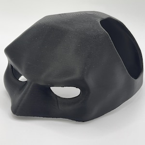 Puede incluir: Una máscara de plástico negra para mascarada con agujeros para los ojos y un diseño en relieve. La máscara está inclinada hacia la izquierda.