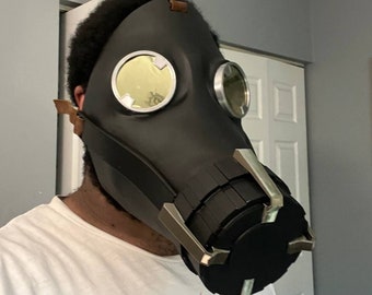 May include: Black gas mask with circular lenses and metallic accents. The mask has brown leather straps and a textured design. The lenses have a gold tint. The mask is worn by a person wearing a white t-shirt.