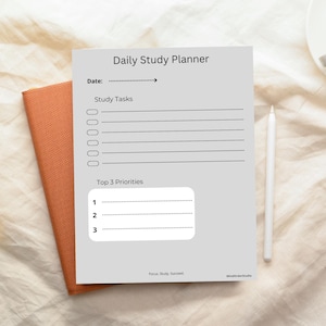 Peut inclure: Un planificateur d'étude quotidien gris avec le texte "Daily Study Planner", "Date:" et "Study Tasks". Le planificateur comprend des sections pour les tâches d'étude et les principales priorités. Un stylo blanc repose à côté du planificateur.