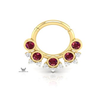 Peut inclure: Un anneau de septum doré avec un design circulaire. Il présente six pierres précieuses rondes, rouge foncé, chacune sertie dans une lunette dorée. De petites pierres blanches sont regroupées autour des gemmes rouges.